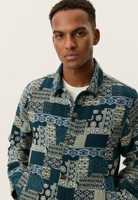 Πουκάμισο πατςγουορκ από φανέλα σε τυρκουάζ και ελιά με ποικίλα σχέδια, κουμπιά στο μπροστινό μέρος, μυτερός γιακάς και υφή υφάσματος.