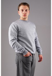 Grauer Pullover mit gerippten Bündchen und Halsausschnitt, mit einem kleinen Emblem auf der Brust. Getragen mit dunkelgrauen Jeans, die Hände in den Taschen.