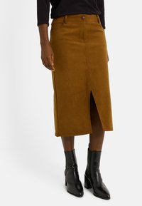 Jupe midi en velours côtelé marron avec une fente avant, équipée de cinq poches et d'une fermeture à boutons. Associée à des bottines noires à talons.