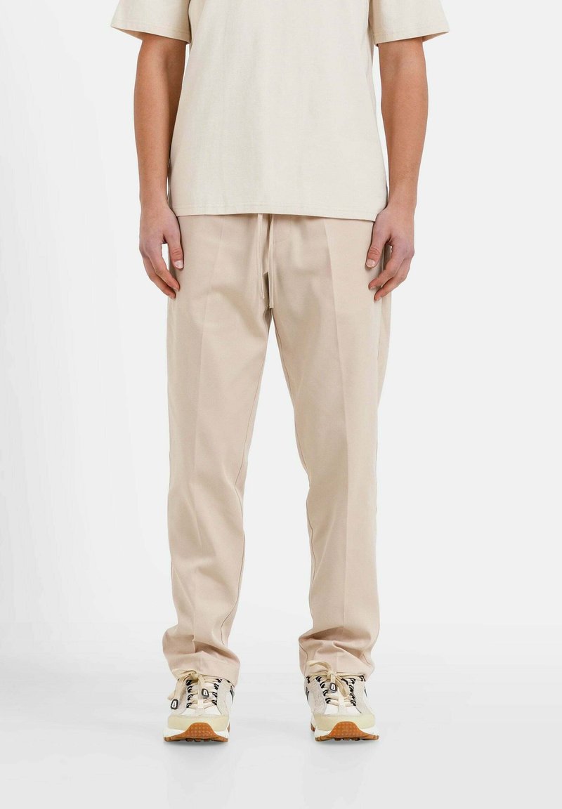 Beige, gefaltete Hose aus weichem Stoff, kombiniert mit einem hellen T-Shirt und beigen Sneakers mit schwarzen Akzenten und einer strukturierten Sohle.