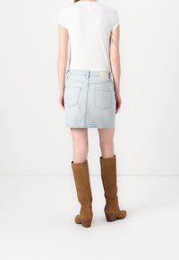 Femme portant une chemise blanche à manches courtes, une jupe en jean bleu clair à motifs et des bottes marron montant jusqu’aux genoux, vue de dos.