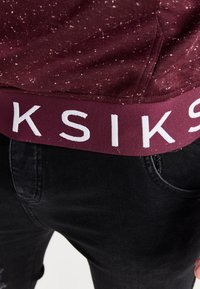 Persona che indossa jeans neri e una cintura color bordeaux con lettere bianche su una maglietta a pois bordeaux visibile sopra la cintura.