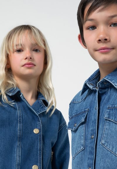 Due bambini indossano giubbotti di jeans blu con cuciture visibili, chiusure a bottoni e taschini sul petto. La texture appare robusta e informale.