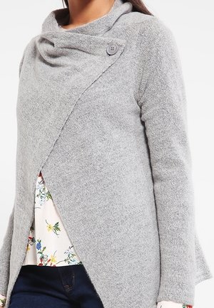 Vrouw draagt een zachte grijze gebreide cardigan met een asymmetrische knoopkraag over een bloemenblouse en donkere spijkerbroek.