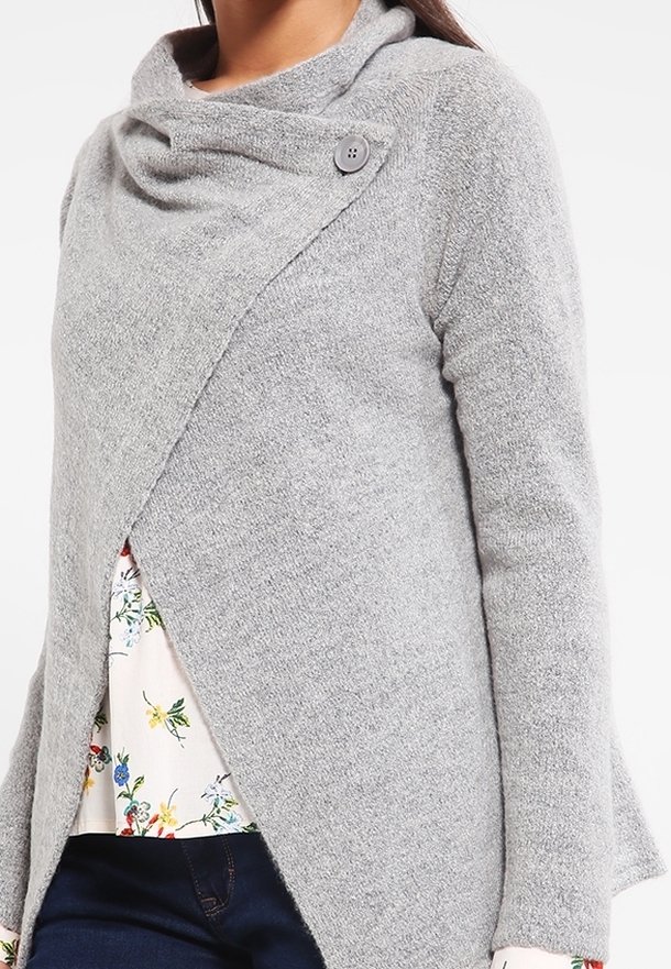 Mujer que lleva un cárdigan de punto gris suave con un cuello abotonado asimétrico sobre una blusa floral y jeans oscuros.