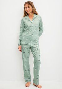 Woman standing barefoot wearing sage green pajamas with a white dog pattern, button-up top, and matching loose pants against a white background.