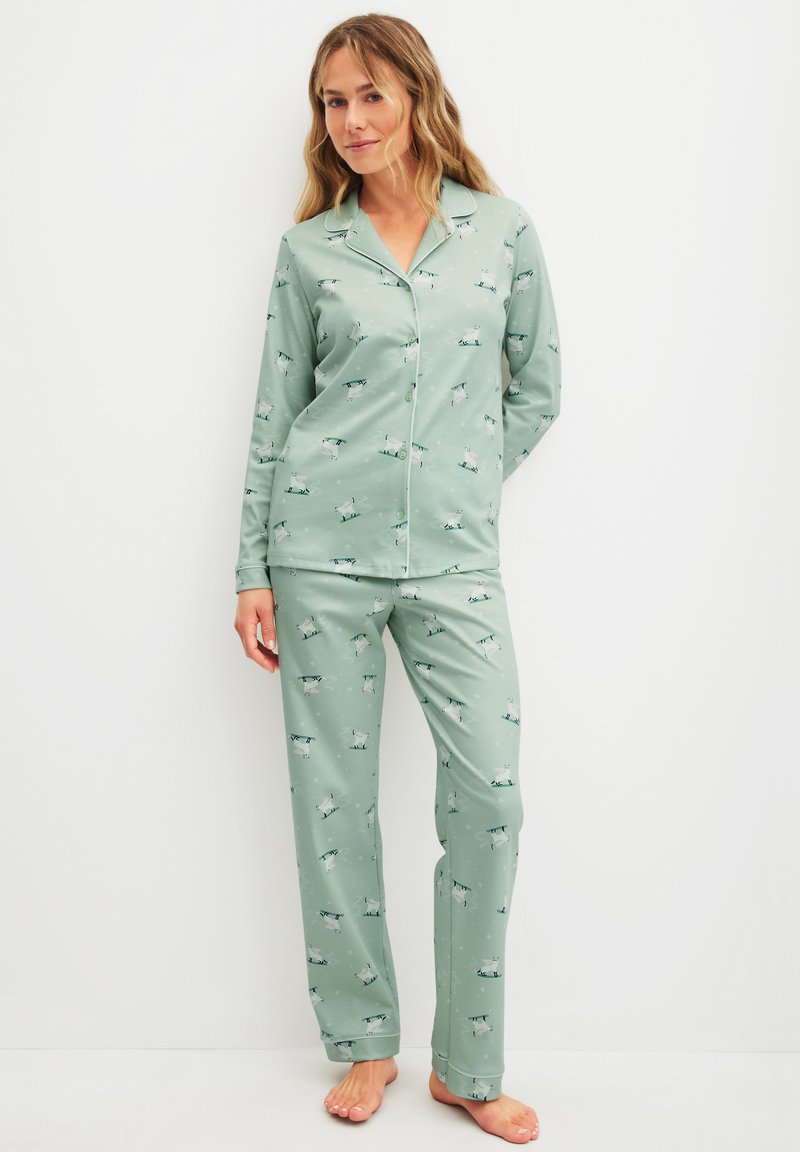 Woman standing barefoot wearing sage green pajamas with a white dog pattern, button-up top, and matching loose pants against a white background.