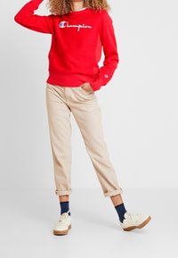 Rode katoenen sweatshirt met geborduurd "Champion" logo, beige relaxte fit broek, witte sneakers met bruine zolen en marineblauwe sokken.