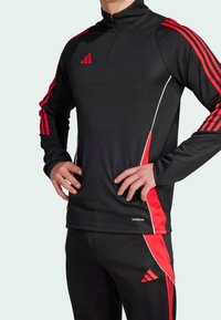 Persona vestida con ropa deportiva negra de Adidas con franjas rojas, de pie con las manos en las caderas, mostrando la marca Aeroready en la parte superior.