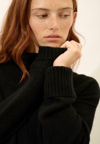 Czarny sweter z dzianiny z wysokim, prążkowanym kołnierzem i grubymi, prążkowanymi mankietami. Miękka tekstura, zbliżenie na ręce i dekolt.