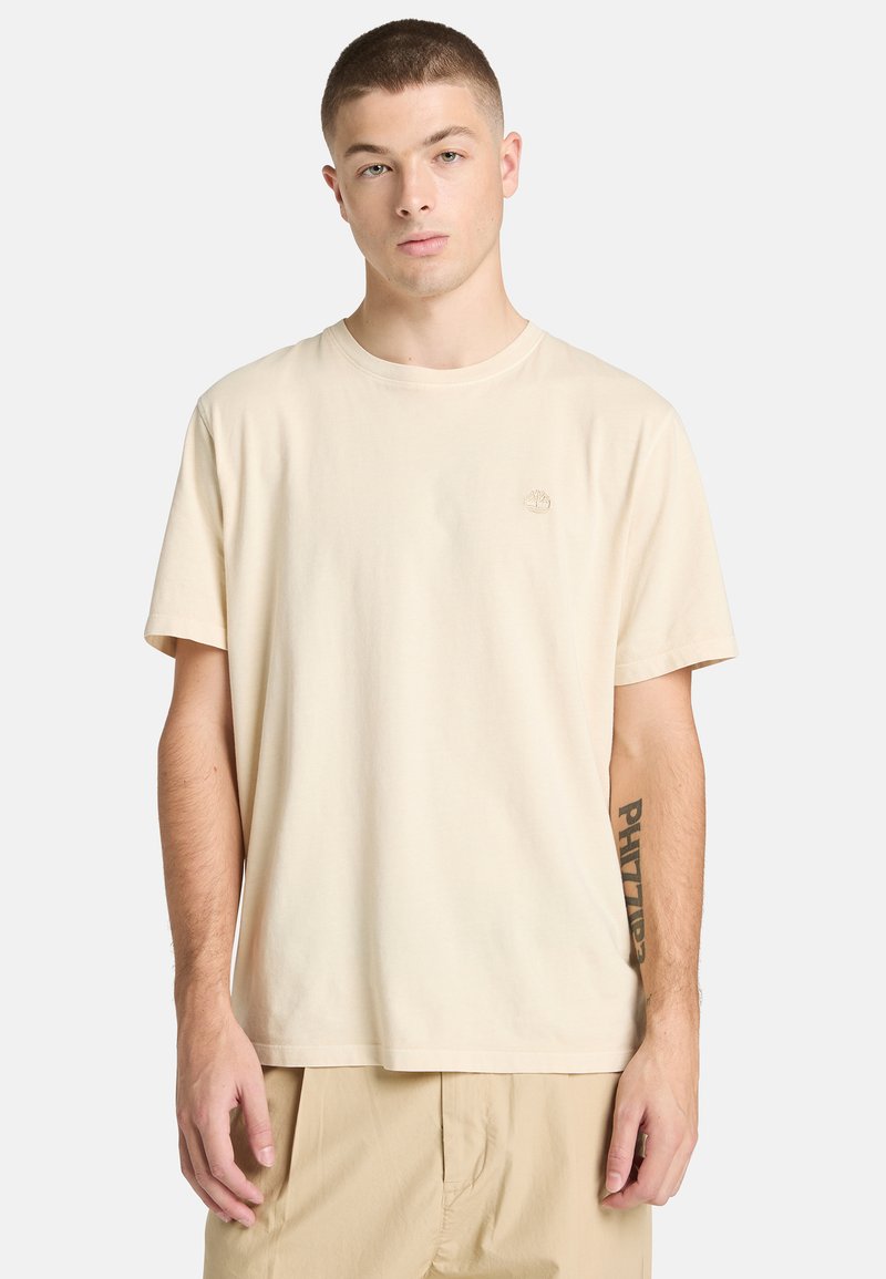 T-shirt en coton beige avec un col rond classique, des manches courtes et une broderie de logo discrète sur la poitrine. Texture lisse, coupe décontractée.