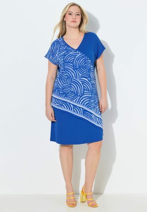 Mujer con vestido azul de manga corta con patrón abstracto de remolinos blancos y sandalias amarillas de tacón, de pie contra un fondo claro y liso.