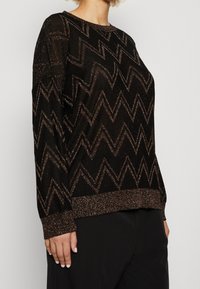 Pull noir avec un motif zigzag brillant, doté de manches longues et d'un poignet côtelé. Le tissu présente une apparence douce et texturée.