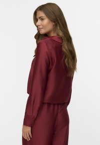 Burgunderfarbige, strukturierte Bluse mit langen Ärmeln und Kragen, mit geknöpften Manschetten und lockerer Passform, kombiniert mit passenden Hosen.