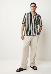 Camisa de manga corta con botones y patrón de rayas blancas y negras, combinada con pantalones de color beige claro y sandalias negras. Textura de tela suave.