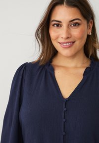 Blusa azul marino con una tela texturizada, que presenta un escote en V, un cuello con volantes y cinco botones delanteros. Mangas suaves y fluidas.