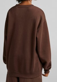 Brun oversized sweatshirt tillverkad av mjukt tyg, med rund halsringning och ribbade muddarna. Baksidan är slät utan synliga logotyper.