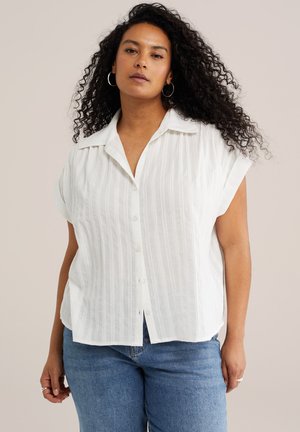 Käharate juustega naine, kes kannab valget lühikeste varrukatega nööpidega särki ja siniseid teksaseid, seisab ühevärvilise tausta ees.