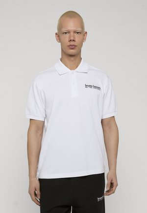 Persona joven con cabello corto y rubio, vistiendo una camisa polo blanca y pantalones negros, ambos con el logo de "bruno banani" en el pecho y el muslo.