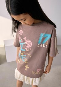 Hnědý oversize tričko s modrou a růžovou grafikou, zdobené květinovými vzory a textem "17 CALI". V kombinaci s bílou krajkovou sukní.