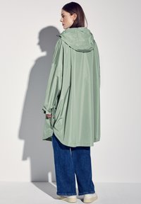 Jasnozielony, wodoodporny płaszcz przeciwdeszczowy z kapturem, zwężoną w talii i szerokimi rękawami. W zestawie z ciemnoniebieskimi spodniami o szerokich nogawkach i kremowymi butami.