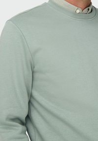 Felpa verde chiaro con colletto a coste, dalla texture morbida e dal fit rilassato. Indossata con una camicia con colletto sotto.