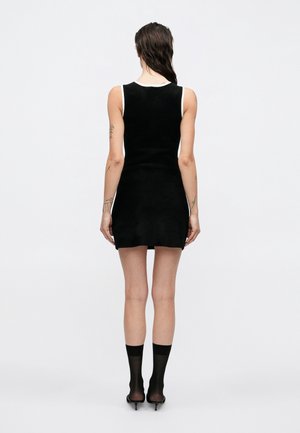 Vrouw met nat haar, gekleed in een mouwloze zwarte minijurk met witte bies, doorschijnende zwarte sokken en zwarte sandalen met hoge hak, staand met de rug naar de camera.