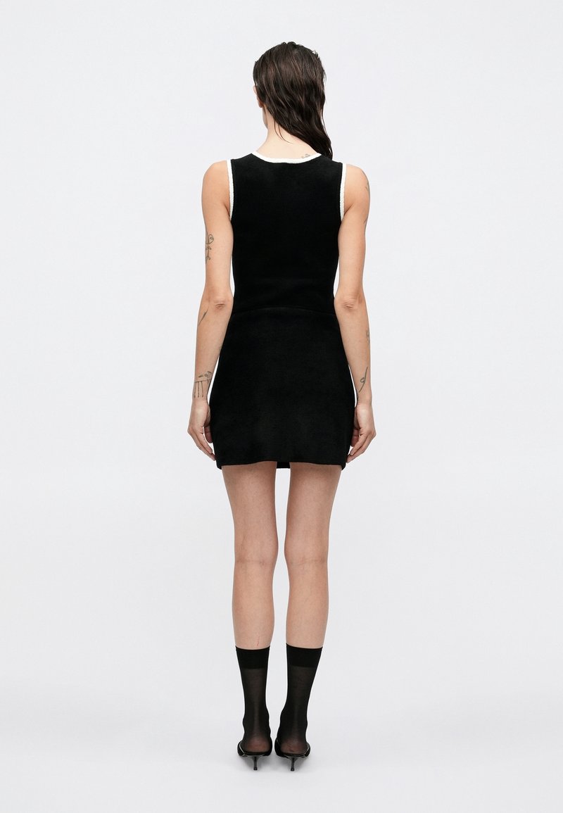 Femme aux cheveux mouillés portant une mini robe noire sans manches avec un liseré blanc, des chaussettes noires transparentes et des sandales à talons hauts noires, debout de dos.