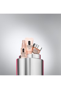 Trzy produkty do pielęgnacji skóry Clinique na srebrnym piedestale: spray, tubka i słoiczek, wszystkie w jasnoróżowym opakowaniu z czarnym nadrukiem.