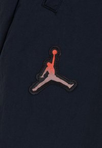Čierna látková nášivka s červeným a šedým logo Jumpman, zobrazujúca siluetu basketbalistu v skákajúcej póze.