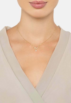 Gold necklace featuring a wavy design with a small diamond accent, resting on a neutral-colored fabric top, highlighting its refined shape.