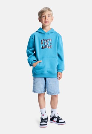 Blauwe trui met capuchon, voorzien van een grafisch logo aan de voorkant, gecombineerd met spijkershorts en zwart-witte-rode sneakers.