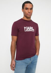 Muž v burgundském tričku s krátkým rukávem a bílým logem "KARL LAGERFELD", v kombinaci s modrými džíny, stojící na jednoduchém bílém pozadí.