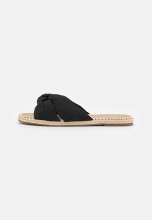 Flad sandal med vævet beige sål og bred sort stofrem snoet over toppen, sidevisning på hvid baggrund.