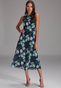 Blomstret midikjole i mørk blå med hvite og grønne blomster mønstre. Ermeløs design med en tettsittende overdel og flytende skjørt. Sølvfargede hæler.