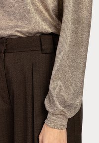 Arm in een lange mouw beige geribbelde top, gecombineerd met hooggewaisted donkerbruine visgraatbroek met riemlussen.