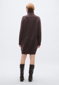 Καφέ πλεκτό φαρδύ φόρεμα με ψηλό γιακά και ριμπ λεπτομέρειες στα μανίκια, συνδυασμένο με μαύρες δερμάτινες μπότες μέχρι το γόνατο με μεταλλικά στοιχεία.