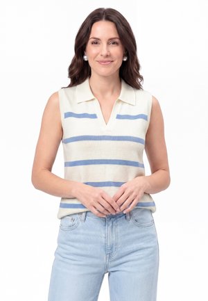 Donna con capelli scuri che indossa orecchini di perle, una canotta bianca con strisce orizzontali blu e jeans azzurri, in piedi con le mani intrecciate.
