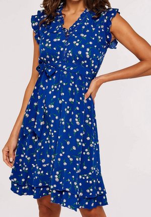 Robe bleue sans manches avec imprimé marguerites blanches, épaules volantées, devant boutonné et ceinture nouée à la taille portée par une femme.
