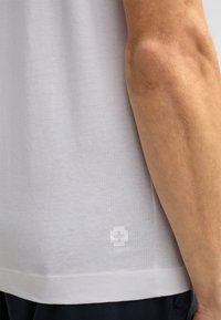 Weißes Baumwoll-T-Shirt mit kurzen Ärmeln, das am Saum ein strukturiertes Logo-Detail aufweist und eine glatte, weiche Stoffoberfläche hat.