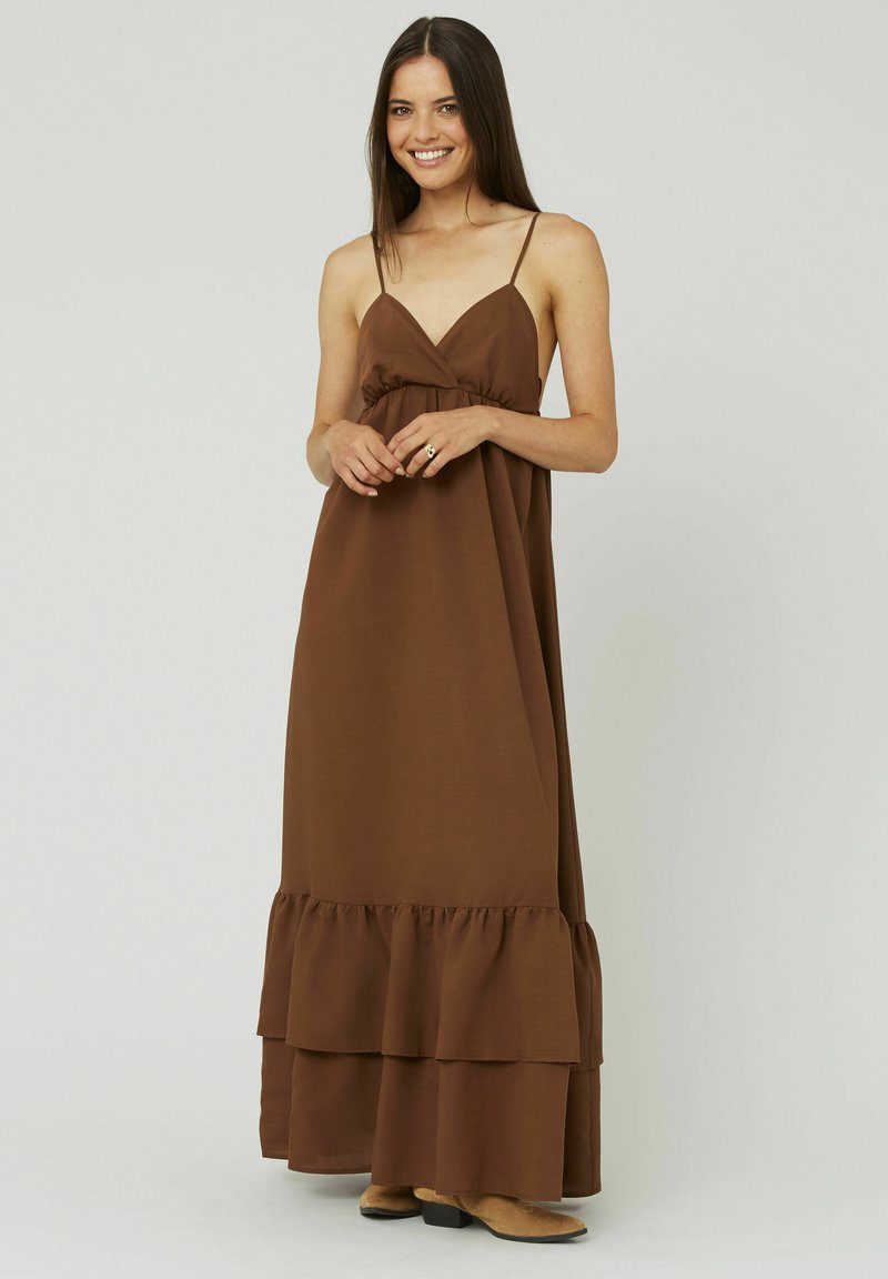 Robe maxi marron avec des bretelles fines, une taille froncée et un ourlet en volants superposés. Tissu lisse avec une finition mate, assorti à des bottes marron clair.