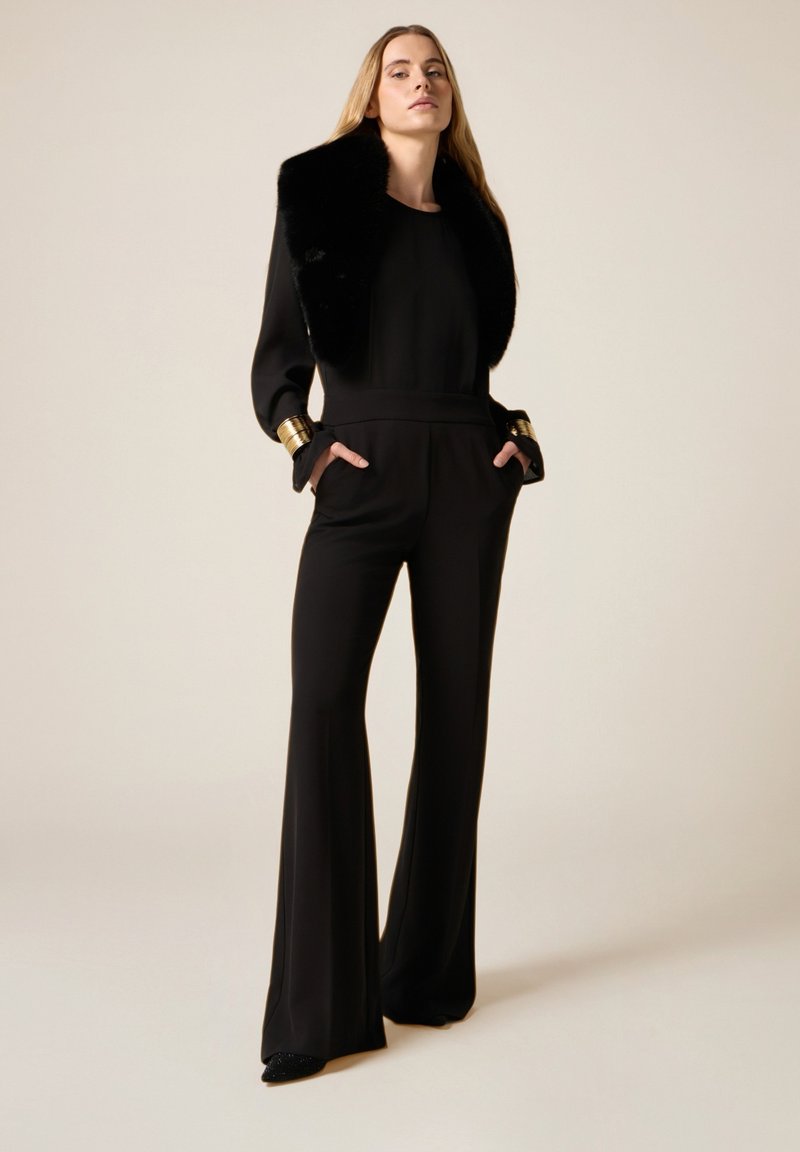 Musta haalari, jossa on istuva yläosa ja levenevät lahkeet, koristeltu tekoturkiskauluksella ja kultaisilla yksityiskohdilla hihansuissa. Sileä rakenne, minimalistinen muotoilu.