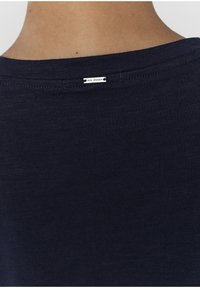 Námorně modré tričko s jemnou, texturovanou tkaninou. Má kulatý výstřih a v zadu je štítek s nápisem "IRIS WOMEN".