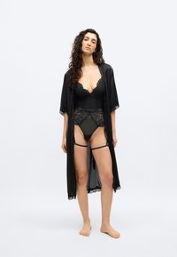 Γυναίκα με μακριά σγουρά μαλλιά στέκεται ξυπόλυτη, φορώντας μαύρα δαντελένια εσώρουχα και ένα ασορτί σατέν ρόμπα με δαντελένιο τελείωμα, μπροστά σε απλό λευκό φόντο.