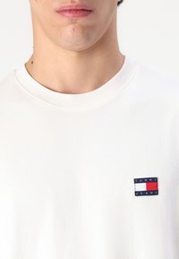 Biely bavlnený tričko s okrúhlym výstrihom a malým obdĺžnikovým logom s červenými, bielymi a modrými blokmi na ľavej hrudi.