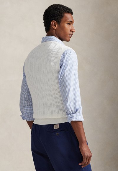 Λευκό γιλέκο με πλέξη καλωδίου πάνω από μπλε πουκαμίσα με καρό, εμφανίζοντας ριμπ τελείωμα και υφή πλέξης. Στο κάτω μέρος φαίνονται σκούρα ναυτικά παντελόνια.