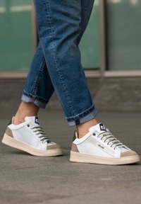 Sneakers bianchi con pannelli in rete e accenti beige, dotati di lacci verde oliva e una suola texture, indossati con jeans blu risvoltati.