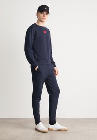 Felpa in pile blu navy con patch rosso, abbinata a pantaloni jogger tapered coordinati. Indossata con sneakers bianche, presenta un cappellino nero.