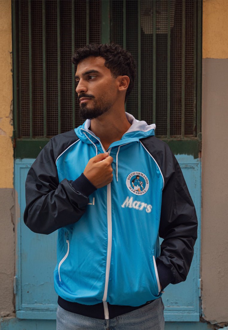 Lichtblauwe en zwarte windjack met rits, logo op de borst, witte accenten en elastische manchetten. Draag met blauwe jeans.