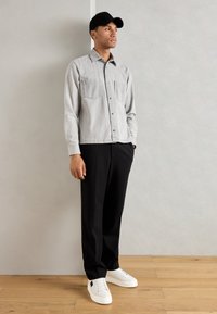 Chemise grise à boutonnières avec deux poches poitrine, pantalon noir et chaussures de sport blanches. Un homme se tient contre un fond uni, portant une casquette noire.